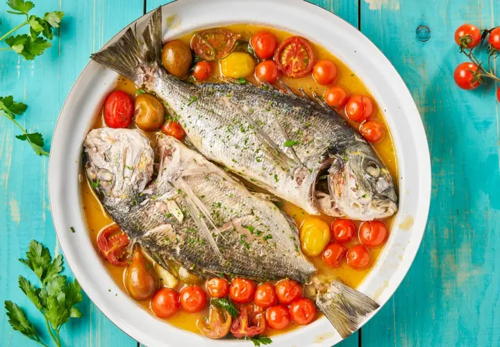 Orata all'acqua pazza, due pesci interi cotti in un brodo saporito con pomodorini e erbe aromatiche, serviti in un piatto bianco su un tavolo di legno blu.