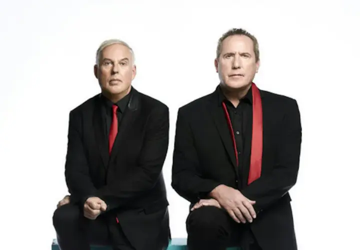Andy McCluskey i Paul Humphreys z OMD, duet znany z niezapomnianych piosenek, w czarnych garniturach i czerwonych akcentach.