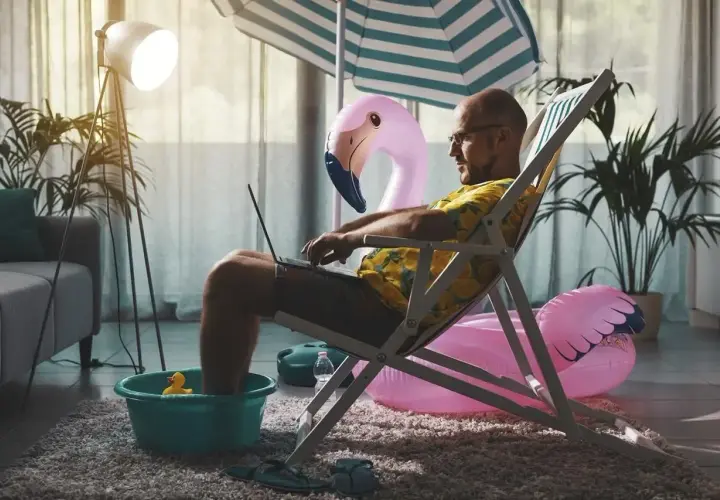 Mężczyzna w hawajskiej koszuli pracuje na laptopie, siedząc w leżaku z nogami w misce. W tle dmuchany flaming i parasol plażowy. Czas wykorzystać zaległy urlop!