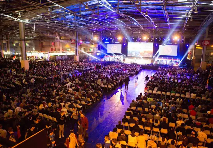 Gran multitud en un evento de convencion, sentada en sillas frente a pantallas gigantes con luces de escenario.