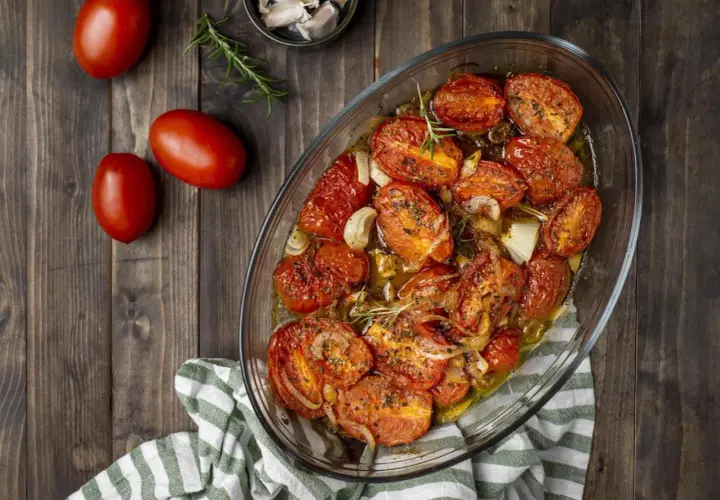 Pomodori gratinati al forno, dorati e succosi, con cipolle e rosmarino in una teglia di vetro su un tavolo di legno.