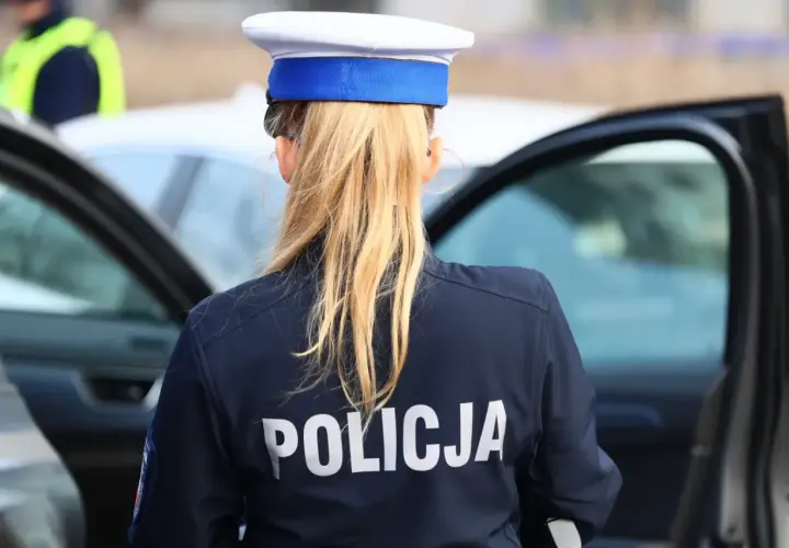 Policjantka w mundurze z napisem "POLICJA" na plecach, stojąca obok otwartych drzwi radiowozu. To jej dodatek funkcyjny.