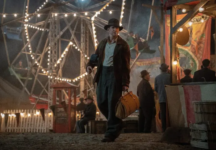 Un uomo con un cappello cammina in una fiera delle illusioni, con una ruota panoramica illuminata sullo sfondo. Le recensioni di Nightmare Alley descrivono questo mondo oscuro.