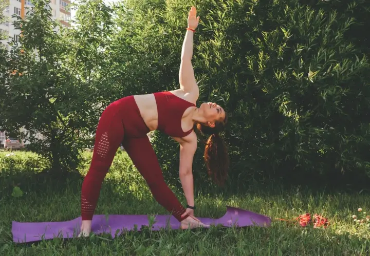 Kobieta w czerwonym stroju wykonuje pozycję Trójkąta podczas praktyki jogi ashtanga na trawie, otoczona zielenią.