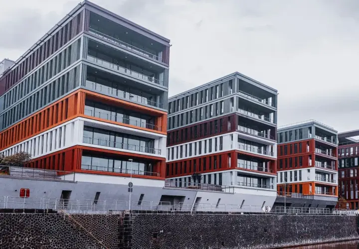 Nowoczesne budynki o geometrycznych kształtach, z elementami w kolorze pomarańczowym i bordowym, tworzą futurystyczny krajobraz miasta nad wodą.