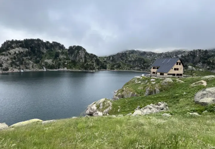 Refugio de montaña junto a un lago rodeado de montañas rocosas y vegetación.