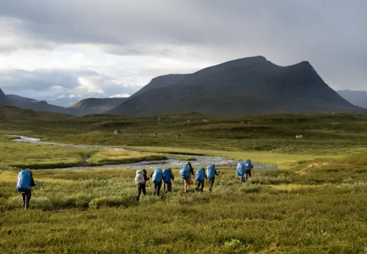 Hikers with blue backpacks trek through a vast, green landscape in Sweden, reminiscent of the Fjällräven Classic. Tents dot the distant hills under a dramatic sky.
