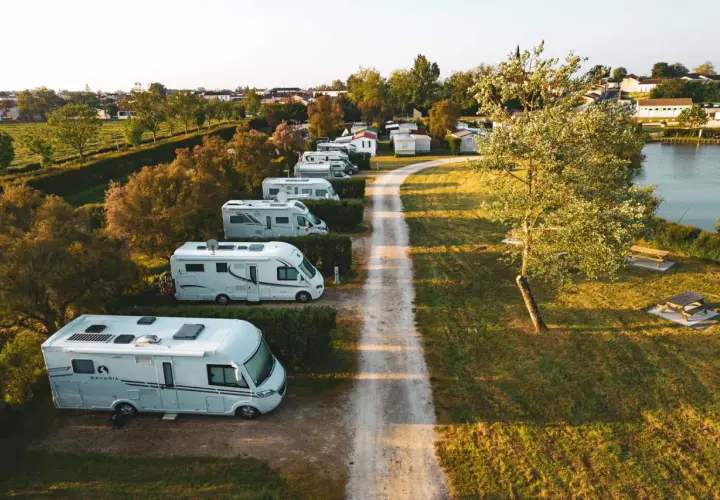 Vue aérienne d'un air camping car park, avec plusieurs camping-cars blancs alignés le long d'un chemin de terre, près d'un lac et d'une petite ville.