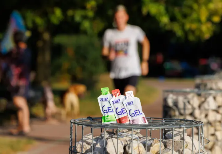 Trzy opakowania żeli energetycznych dla biegaczy: zielone jabłko, cola i truskawka z bananem, na tle biegnącej osoby.