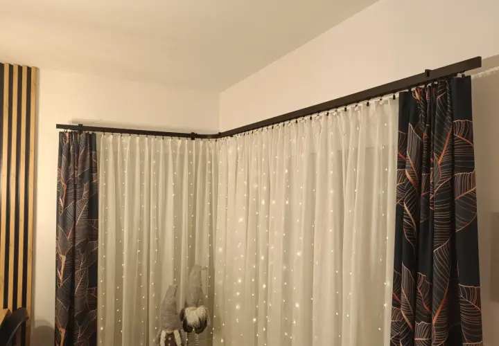 Elegancki karnisz apartamentowy czarny, na którym wiszą zasłony z motywem liści i delikatne firanki ze światełkami.