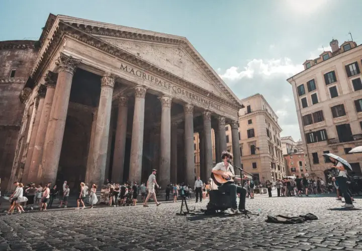 Un musicien joue de la guitare devant le Panthéon d'Agrippa, un monument historique de Rome, tandis que des touristes se promènent sur la place pavée.