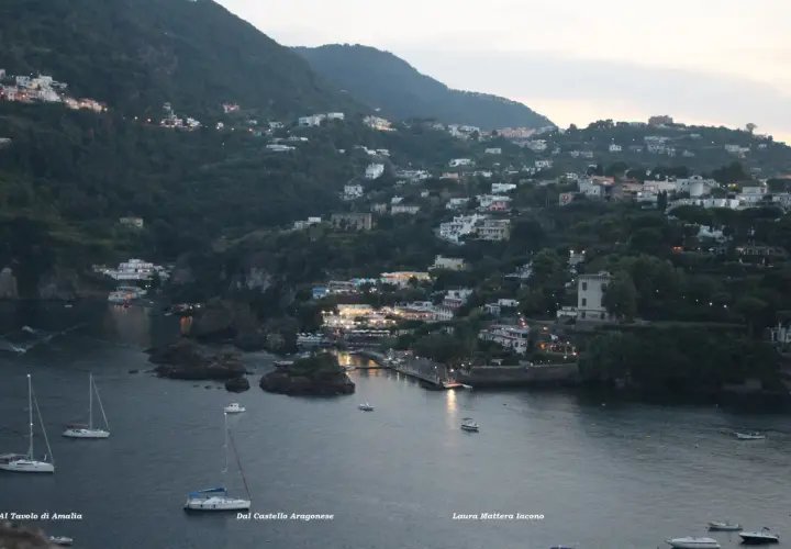 Vittoria Colonna a Ischia: case aggrappate alla collina, luci soffuse e barche a vela nella baia al crepuscolo.