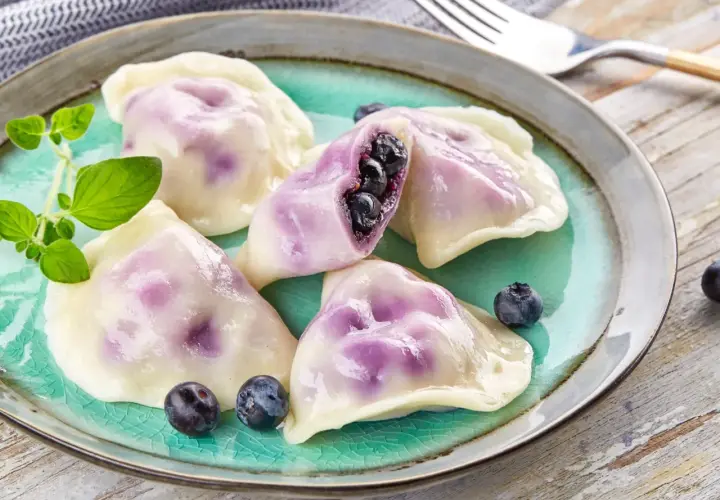 Pyszne pierogi z jagodami, jeden przekrojony, ukazujący soczyste owoce. Podane na turkusowym talerzu z gałązką ziół.