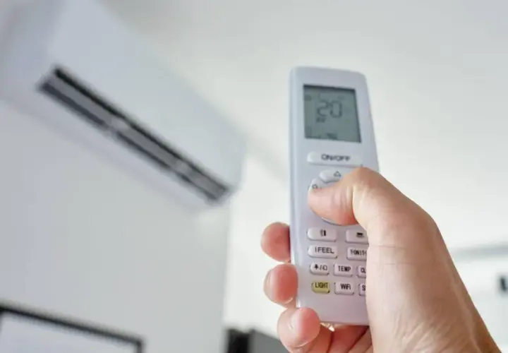 Mano sujetando un control remoto para encender aire acondicionado. La unidad interior está en el fondo.