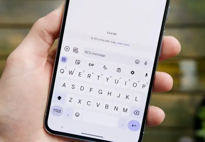 Dłoń trzyma smartfon z włączoną klawiaturą. Na ekranie widoczny jest czat RCS z Judy.