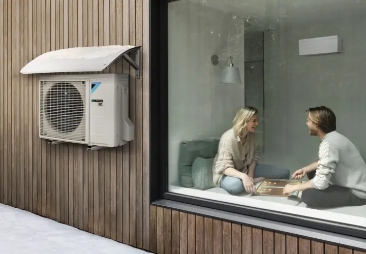 Unidad exterior de aire acondicionado Daikin instalada con la distancia adecuada a la pared de madera. Dentro, una pareja juega.