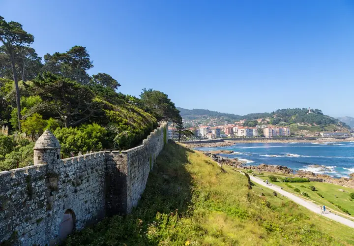 A historic stone wall winds along the coast of Baiona, Portugal, overlooking the blue sea and a town nestled on the hills.