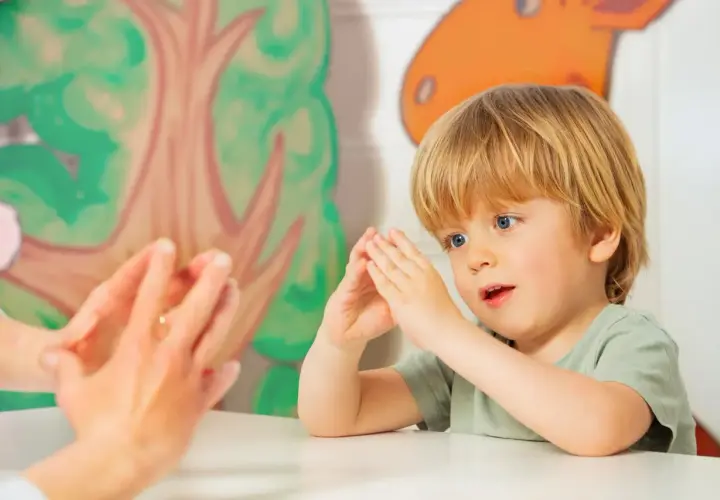 Ein Junge mit blonden Haaren und blauen Augen macht Fingerspiele im Kindergarten. Er schaut gespannt zu, wie die Hände einer Erwachsenen vor ihm tanzen.