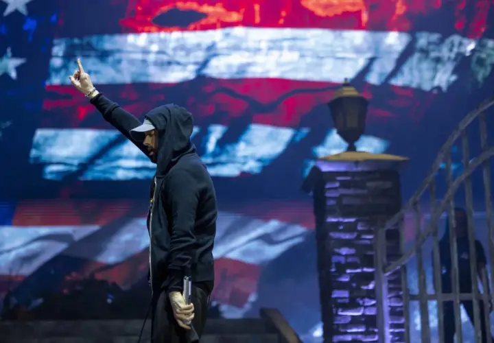 Eminem na koncercie w Polsce, z ręką uniesioną w geście pozdrowienia, na tle ekranu z amerykańską flagą.