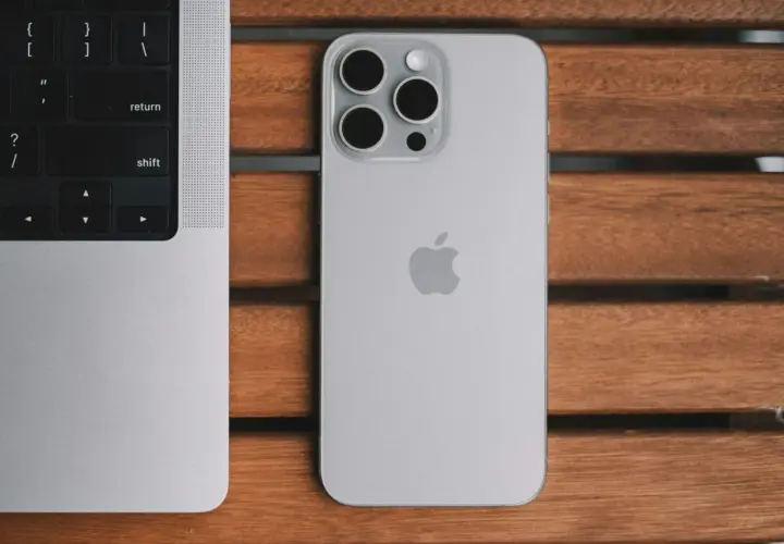 Srebrny iPhone 13 Pro z systemem iOS 26.4 obok klawiatury MacBook'a na drewnianym stole.