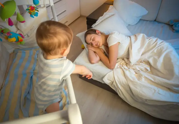 Un neonato di 7 mesi, in piedi nella sua culla, osserva la mamma addormentata nel letto.