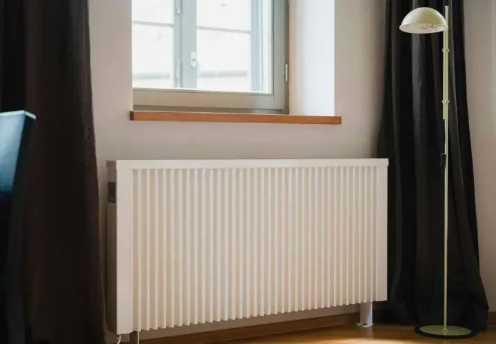 Biały kaloryfer pod oknem, obok ciemnej zasłony i lampy stojącej. Dowiedz się, jak ustawić kaloryfery przy ogrzewaniu gazowym dla optymalnego komfortu.