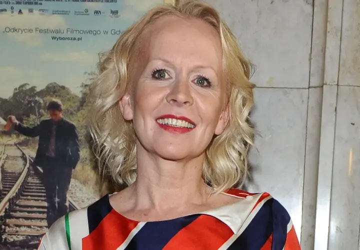 Agnieszka Krukówna uśmiecha się, ubrana w kolorowy strój. W tle plakat filmowy z parą na torach.
