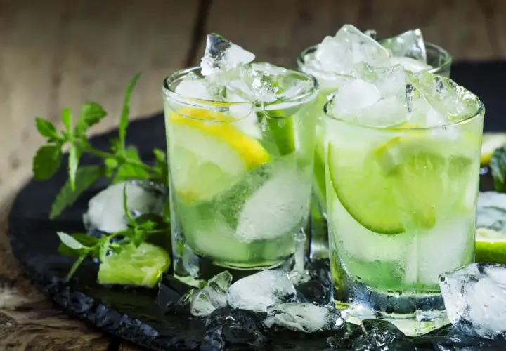Trois verres de caipirinha glacée, garnie de citron vert et de citron, sur un plateau noir avec de la menthe fraîche. La recette parfaite pour se rafraîchir.