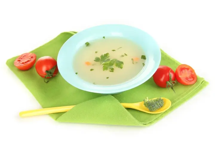 Brodo vegetale neonati in ciotola azzurra, con pomodorini e un cucchiaino giallo con broccoli.