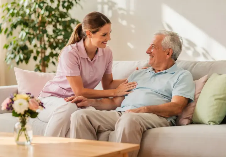 Eine Pflegerin spricht lächelnd mit einem älteren Mann auf einem Sofa. Ein Gespräch über den Eigenanteil im Pflegeheim kann so angenehm sein.