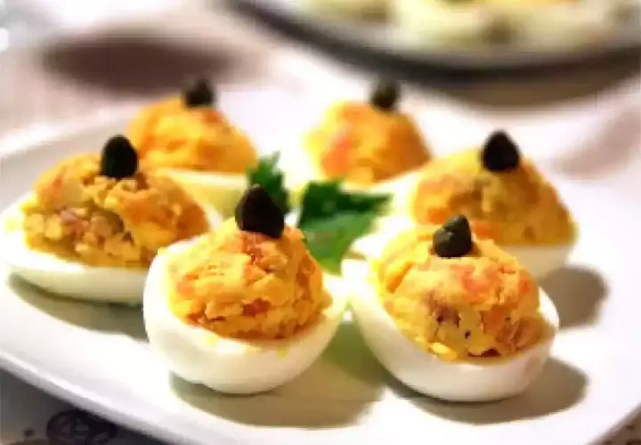 Six oeufs mimosa saumon garnis d'une olive noire sur une assiette blanche.
