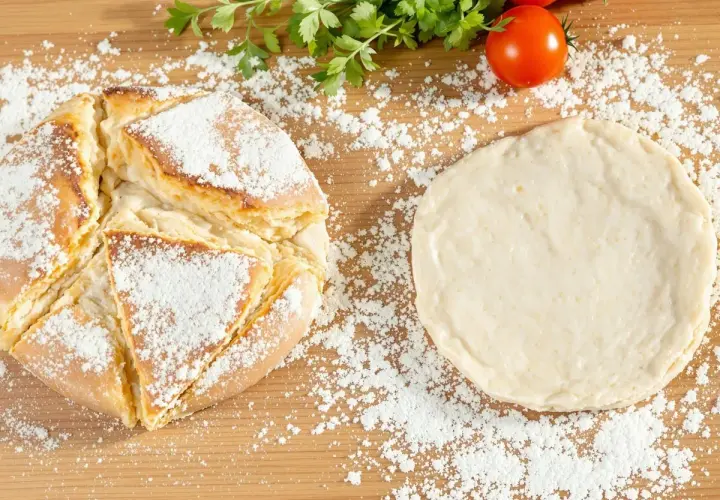 Une recette de pain maison : une miche dorée saupoudrée de farine à côté d'une pâte à pain crue, avec des tomates et du persil en arrière-plan.