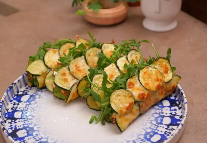 Rotolini di zucchine e rucola, cotti in padella e gratinati con formaggio fuso, serviti su un piatto decorato.