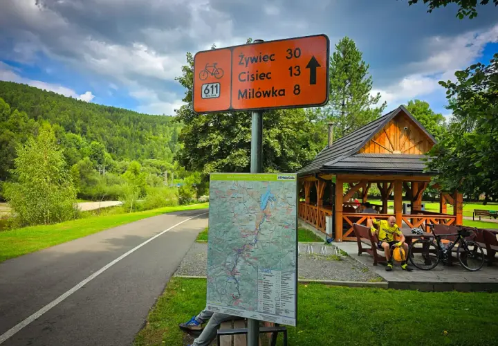 Znak szlaku rowerowego Velo Sola z odległościami do Żywca, Ciśca i Milówki. Obok mapa i wiata.