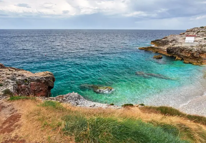 Vue d'une crique aux eaux turquoise cristallines, typique d'une plage de Pula en Sardaigne. Rochers et végétation méditerranéenne bordent la petite plage de galets.