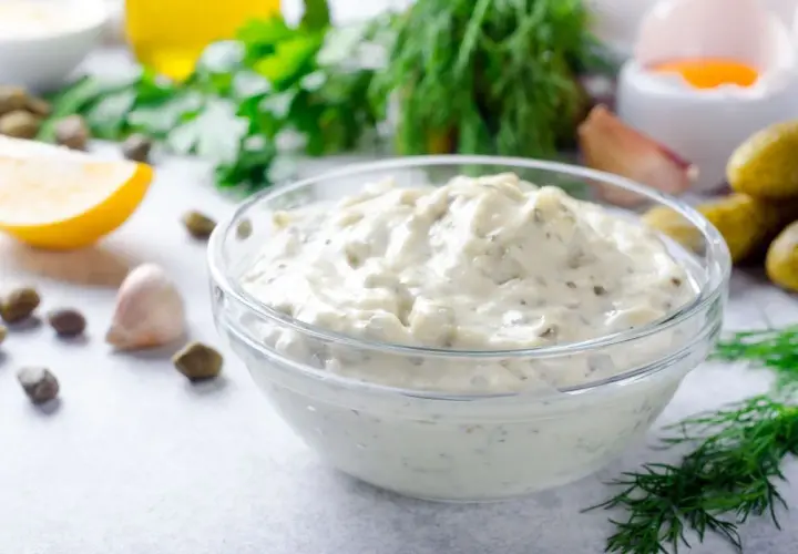 Una ciotola di cremosa salsa tartara, con capperi, limone, aglio e aneto sullo sfondo.