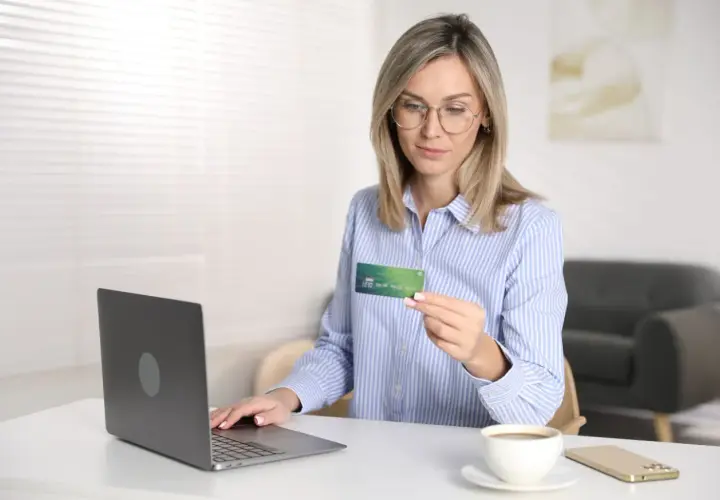 Kobieta z kartą płatniczą w ręku, gotowa do dokonania płatności online. Na biurku laptop, telefon i kawa. Reserved Paypo.
