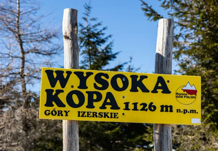 Żółta tablica na dwóch drewnianych słupkach informuje: "Wysoka Kopa 1126 m n.p.m. Góry Izerskie".