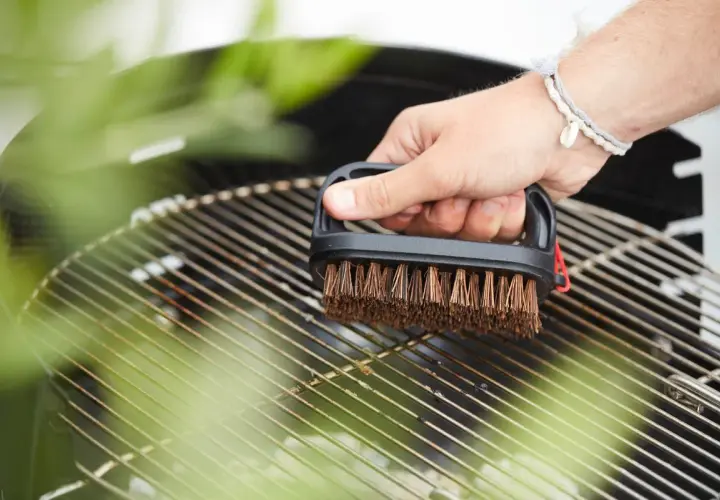 Main tenant une brosse pour nettoyer la grille du barbecue. Les herbes vertes floutées ajoutent une touche de fraîcheur à la scène.