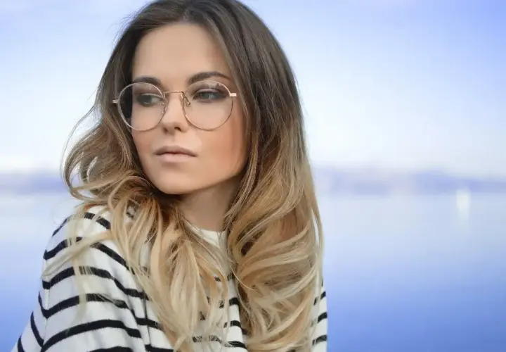 Blondynka w okularach o okrągłych oprawkach, w pasiastym swetrze, patrzy w bok na tle błękitnego nieba i wody.