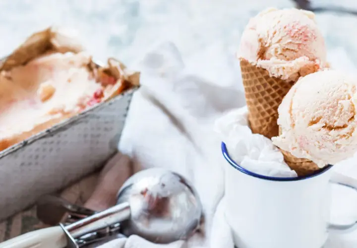 Due palline di gelato fatto in casa, con pezzetti di fragola, in un cono, accanto a una vaschetta e un porzionatore.