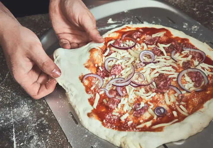 Hände ziehen den rohen Pizzateig vom Pizzaschieber. Der Teig klebt leicht am Schieber, während die Pizza mit Tomatensauce, Käse, Salami und Zwiebeln belegt ist.
