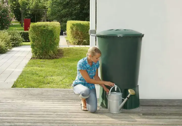 Donna raccoglie acqua piovana con un annaffiatoio da un serbatoio verde in giardino. Un modo ecologico per innaffiare le piante.
