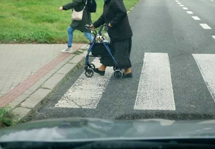 Starsza pani z chodzikiem i młodsza osoba przechodzą przez przejście dla pieszych.
