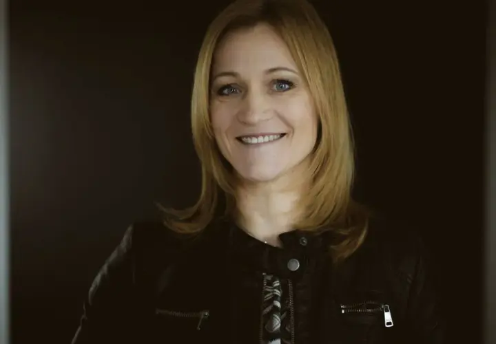 Uśmiechnięta blondynka w czarnej ramonesce na ciemnym tle.