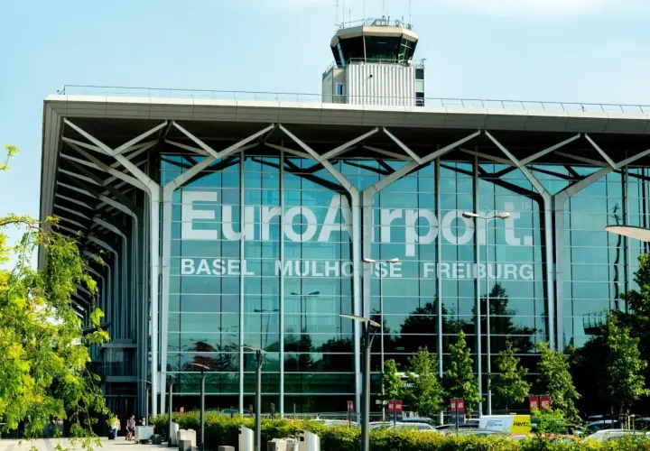Nowoczesny budynek lotniska EuroAirport Basel Mulhouse Freiburg z charakterystyczną wieżą kontroli lotów i zielenią przed fasadą.