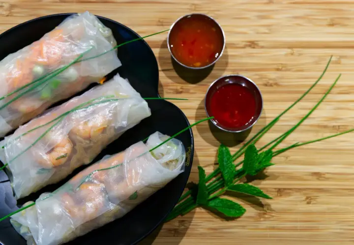 Tres rollitos de verano frescos con camarones y verduras, servidos en un plato negro con dos salsas y hojas de menta.