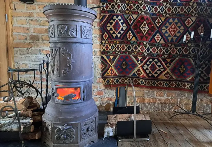 Stary, ozdobny kominek czy koza z płonącym ogniem, obok stos drewna i kolorowy kilim na ceglanej ścianie.