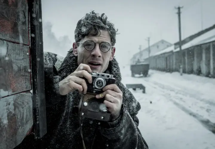 Mężczyzna w okularach z aparatem w ręku, w zimowej scenerii. Scena jak z filmów Agnieszki Holland.