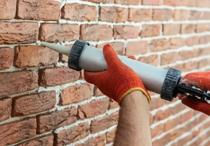 Ręce w pomarańczowych rękawiczkach nakładają masę uszczelniającą na fugę między cegłami, tworząc estetyczne wykończenie.